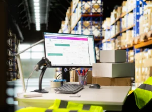 Puesto de trabajo en un almacén con computadora, escáner de códigos de barras y estanterías al fondo.