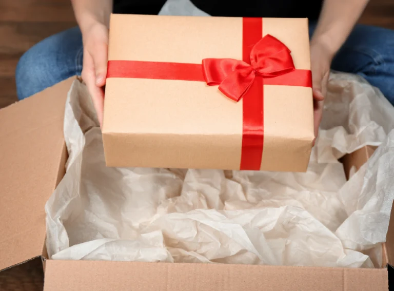 Manos abriendo una caja de cartón con un lazo rojo y papel de seda.