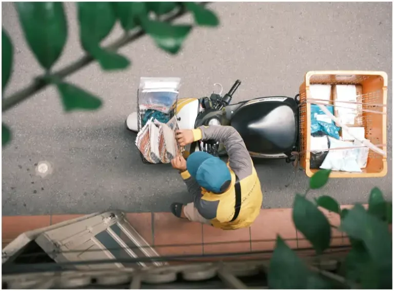 Una vista aérea muestra a un repartidor local cargando periódicos y paquetes en su motocicleta, estacionada en una calle adoquinada junto a una puerta tradicional. Lleva una chaqueta de uniforme amarilla y gris y una gorra de béisbol verde.