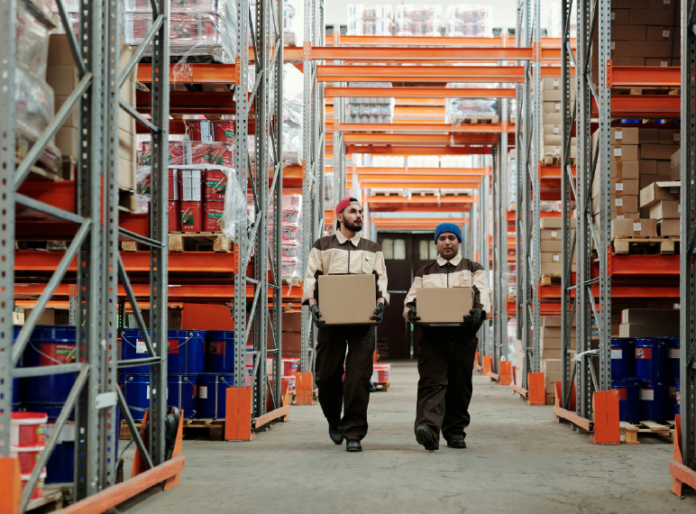 dos  personas caminando con dos cajas en mano en un almacen fulfillment para ver donde van a poner las cajas que tienen cargando
