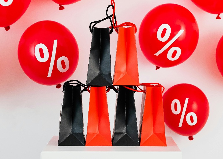 Pirámide de bolsas de regalo negras y naranjas frente a globos rojos con símbolos de porcentaje, representando ofertas de Hot Sale y envíos e-commerce.