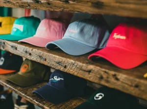 Varias gorras de visera curva de diferentes colores (rosa, azul, roja, verde) acomodadas en estantes de madera rústica de una tienda de ropa o almacén de e-commerce.