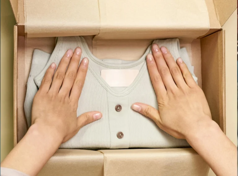 Vista aérea de unas manos acomodando una prenda de ropa acanalada de color verde claro con botones, perfectamente doblada sobre papel kraft dentro de una caja de cartón para envío. Envíos de ropa