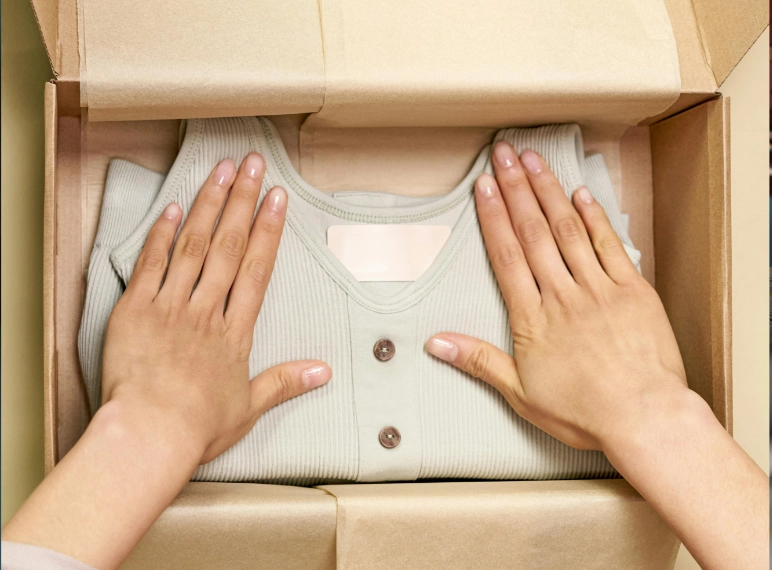 Vista aérea de unas manos acomodando una prenda de ropa acanalada de color verde claro con botones, perfectamente doblada sobre papel kraft dentro de una caja de cartón para envío. Envíos de ropa