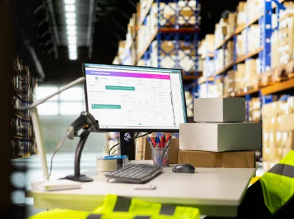 Puesto de trabajo en un almacén con computadora, escáner de códigos de barras y estanterías al fondo.